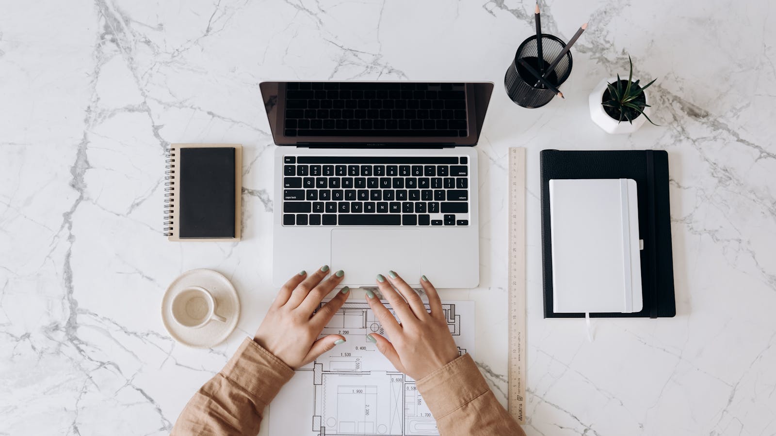 Top view of a stylish home office desk with a laptop, planner, and coffee cup, showing hands on a blueprint. (Website Maintenance) Made Easy: Let Priceless Consulting Handle It